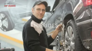  Here is the short alt text for the requested image: A mechanic in a black cap and glasses looks at the camera while adjusting a wheel alignment sensor on a dark SUV.