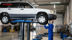 A mechanic in blue overalls stands in an auto repair shop, inspecting the unders ide of a white Subaru Forester SUV that has been raised high on a hydraulic lift.