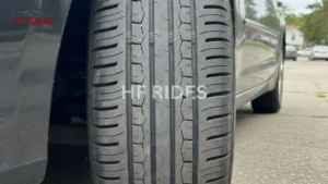 Close-up of a car tire with detailed tread patterns. The vehicle is parked on a paved surface with a suburban neighborhood in the background.